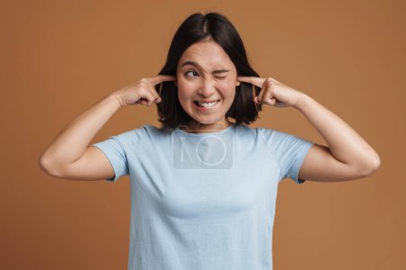 Téléchargez les photos : Jeune femme asiatique portant t-shirt grimacing tout en branchant ses oreilles isolées sur fond beige - en image libre de droit