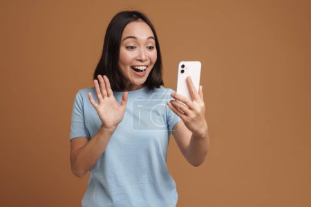 Photo for Excited asian woman wearing t-shirt gesturing while using cellphone isolated over beige background - Royalty Free Image