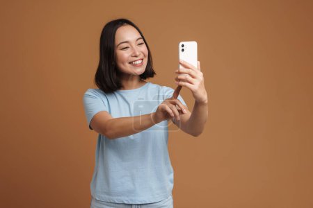 Téléchargez les photos : Jeune femme asiatique souriant tout en prenant selfie sur téléphone portable isolé sur fond beige - en image libre de droit