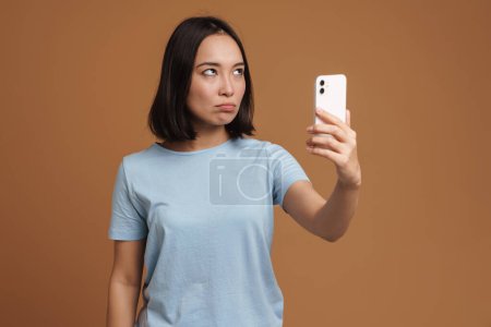Photo for Displeased asian woman taking selfie photo on cellphone isolated over beige background - Royalty Free Image