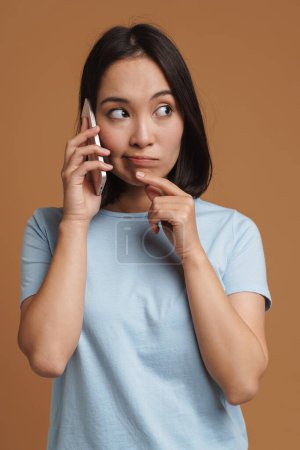 Téléchargez les photos : Femme asiatique perplexe portant t-shirt parler sur téléphone cellulaire isolé sur fond beige - en image libre de droit