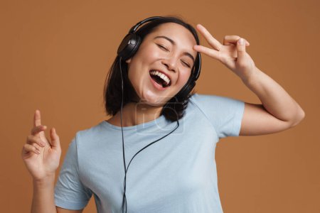 Photo for Young asian woman in headphones dancing while listening music isolated over beige background - Royalty Free Image