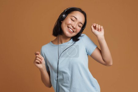 Téléchargez les photos : Jeune femme asiatique dans casque dansant tout en écoutant de la musique isolée sur fond beige - en image libre de droit