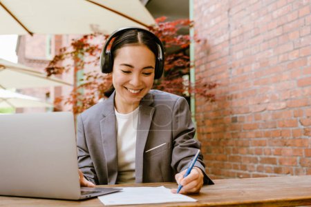 Téléchargez les photos : Femme asiatique dans les écouteurs en écrivant des notes tout en travaillant avec un ordinateur portable dans un café à l'extérieur - en image libre de droit
