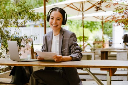 Téléchargez les photos : Femme asiatique dans les écouteurs en écrivant des notes tout en travaillant avec un ordinateur portable dans un café à l'extérieur - en image libre de droit