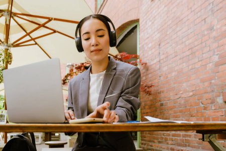 Photo for Asian woman in headphones using gadgets while working at cafe outdoors - Royalty Free Image
