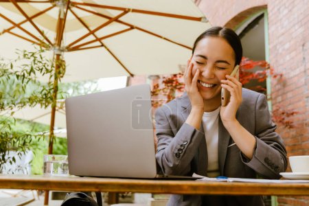 Photo for Asian woman talking on cellphone while working with laptop at cafe outdoors - Royalty Free Image