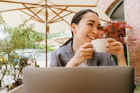 Téléchargez les photos : Femme asiatique buvant du café tout en travaillant avec un ordinateur portable dans un café en plein air - en image libre de droit