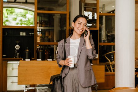 Téléchargez les photos : Asiatique jeune femme d'affaires boire du café et parler sur téléphone portable tout en se tenant dans le café - en image libre de droit