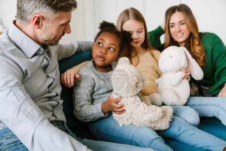 Foto de Familia feliz con la niña africana adoptada sentada en el sofá en la sala de estar en casa. Concepto de adopción. - Imagen libre de derechos