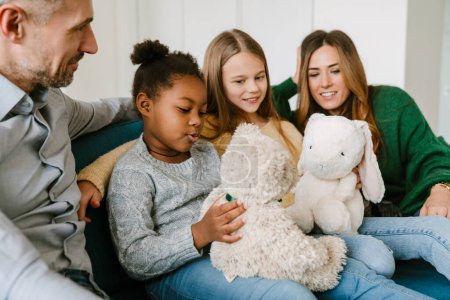 Familia feliz sentada en el sofá, hermanas adoptivas jugando juguetes de peluche. Concepto de adopción.