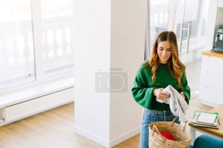 Téléchargez les photos : Belle femme souriante faisant le travail de maison dans la chambre confortable ensoleillée à la maison - en image libre de droit