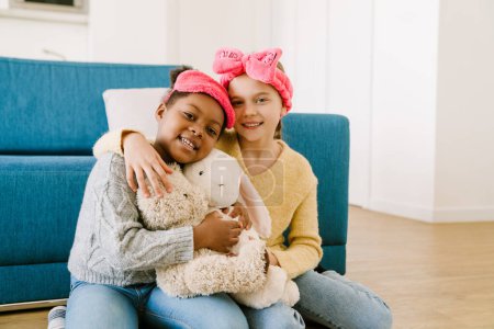 Foto de Poco sonriente feliz interracial hermanas abrazando y jugando con peluches juntos en casa - Imagen libre de derechos