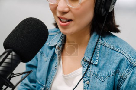 Téléchargez les photos : Belle jeune animatrice de radio heureuse utilisant microphone et écouteurs tout en diffusant en studio - en image libre de droit