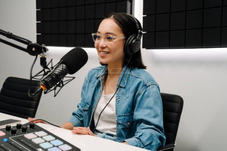 Foto de Hermosa joven feliz anfitrión de radio utilizando micrófono y auriculares mientras se transmite en el estudio - Imagen libre de derechos