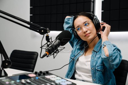 Beautiful happy young female radio host using microphone and headphones while broadcasting in studio