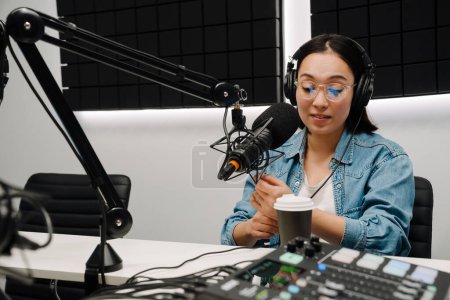 Téléchargez les photos : Belle jeune animatrice de radio heureuse utilisant microphone et écouteurs tout en diffusant en studio - en image libre de droit