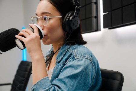 Foto de Joven presentadora de radio femenina con auriculares bebiendo café mientras transmite en estudio - Imagen libre de derechos