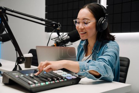 Téléchargez les photos : Belle jeune animatrice de radio heureuse utilisant un microphone et un mixeur sonore tout en diffusant en studio - en image libre de droit