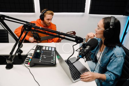 Téléchargez les photos : Jeune homme blanc jouant de la guitare tandis que la femme heureuse enregistrement podcast sur ordinateur portable en studio de radio - en image libre de droit