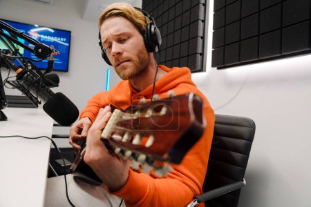 Téléchargez les photos : Jeune homme blanc utilisant microphone et écouteurs tout en jouant de la guitare en studio de radio - en image libre de droit