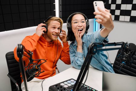 Photo for Happy young man and woman smiling and taking selfie photo while broadcasting in radio studio - Royalty Free Image