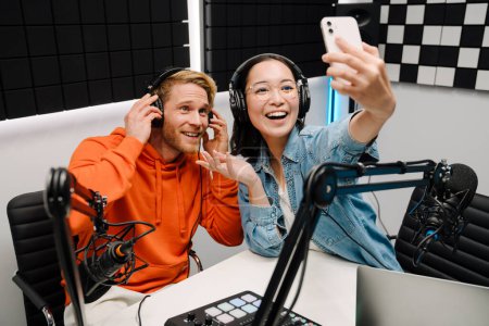 Téléchargez les photos : Joyeux jeune homme et femme souriant et prenant selfie photo tout en diffusant dans un studio de radio - en image libre de droit