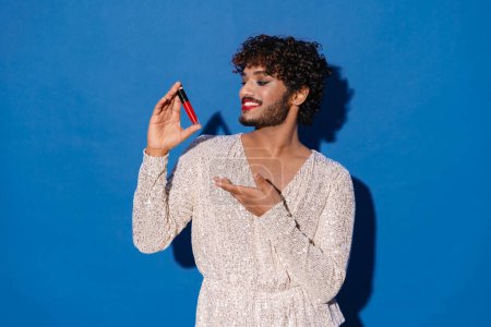 Téléchargez les photos : Jeune homme bouclé avec le maquillage souriant et montrant rouge à lèvres isolé sur fond bleu - en image libre de droit