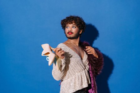 Photo for Young curly man with makeup holding high heels and looking aside isolated over blue background - Royalty Free Image