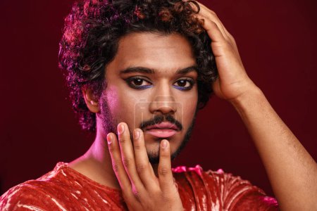Téléchargez les photos : Jeune homme bouclé avec maquillage posant et regardant la caméra isolée sur fond rouge - en image libre de droit