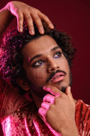 Foto de Joven hombre rizado con maquillaje posando y mirando hacia arriba aislado sobre fondo rojo - Imagen libre de derechos