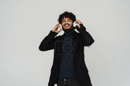 Téléchargez les photos : Jeune adulte indien souriant bouclé homme dans les écouteurs sur fond gris isolé - en image libre de droit