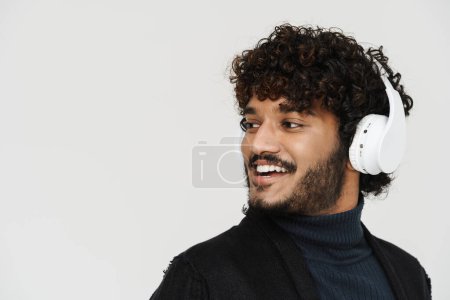 Photo for Adult indian handsome curly man in headphones looking leftwards and smiling over grey isolated background - Royalty Free Image