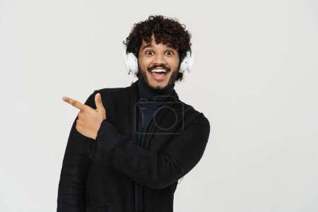 Photo for Adult young indian smiling curly man in headphones pointing leftwards and looking on camera over grey isolated background - Royalty Free Image