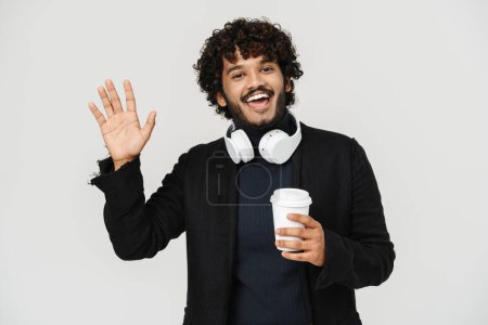 Téléchargez les photos : Jeune adulte indien souriant bouclé homme agitant avec du café regardant la caméra sur fond gris isolé - en image libre de droit