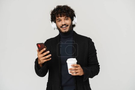 Téléchargez les photos : Jeune adulte indien souriant bouclé homme dans les écouteurs avec café et téléphone regardant la caméra sur fond isolé igrey - en image libre de droit