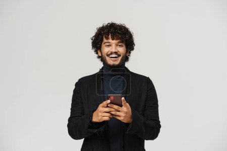 Téléchargez les photos : Jeune homme indien souriant heureux tenant le téléphone et regardant la caméra sur fond gris isolé - en image libre de droit