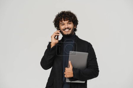 Foto de Joven indio guapo hombre hablando con el teléfono con el ordenador portátil mirando a la cámara en gris aislado fondo - Imagen libre de derechos