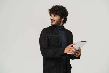 Photo for Adult young indian handsome curly man with tablet looking leftwards and smiling over grey isolated background - Royalty Free Image
