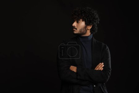 Photo for Young handsome indian curly serious man standing arms folded looking leftwards over isolated dark background - Royalty Free Image