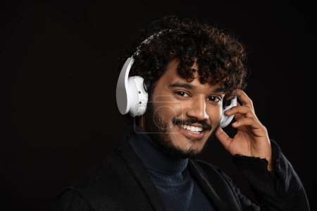 Foto de Retrato de un joven indio rizado sonriente con auriculares mirando a la cámara sobre un fondo oscuro aislado - Imagen libre de derechos