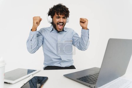 Photo for Young indian glad office employee employee sitting in front of laptop and celebrates with raised arms over grey isolated background - Royalty Free Image