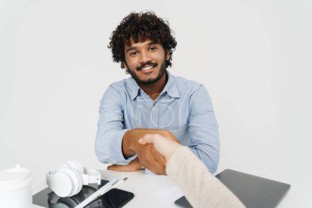 Photo for POV handshake across the table with smiling curly indian man over isolated grey background - Royalty Free Image