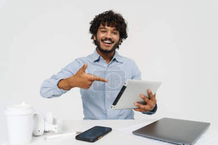 Foto de Feliz hombre indio sonriente sentado en la mesa de la oficina con la tableta y señalándolo sobre el fondo gris aislado - Imagen libre de derechos