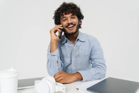 Foto de Joven feliz hombre indio sonriente sentado en la mesa de la oficina y hablando en su teléfono sobre fondo gris aislado - Imagen libre de derechos