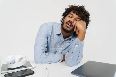 Photo for Adult tired Indian man sleeping at office desk propping his head up over isolated grey background - Royalty Free Image