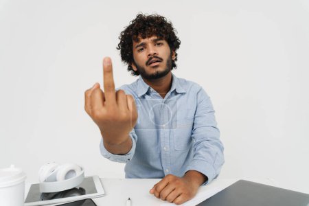 Foto de Empleado de oficina indio adulto está mostrando el dedo medio por la mesa de la oficina con la cara descontenta sobre fondo gris aislado - Imagen libre de derechos
