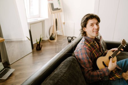 Téléchargez les photos : Homme à poils blancs souriant et jouant ukulélé tout en se reposant sur le canapé à la maison - en image libre de droit