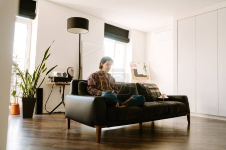 Foto de Hombre blanco usando camisa usando el ordenador portátil mientras descansa en el sofá en casa - Imagen libre de derechos