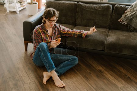 Photo for White man in glasses using mobile phone while sitting on floor at home - Royalty Free Image